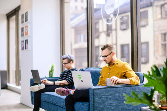 two people sitting on a couch working on laptops
